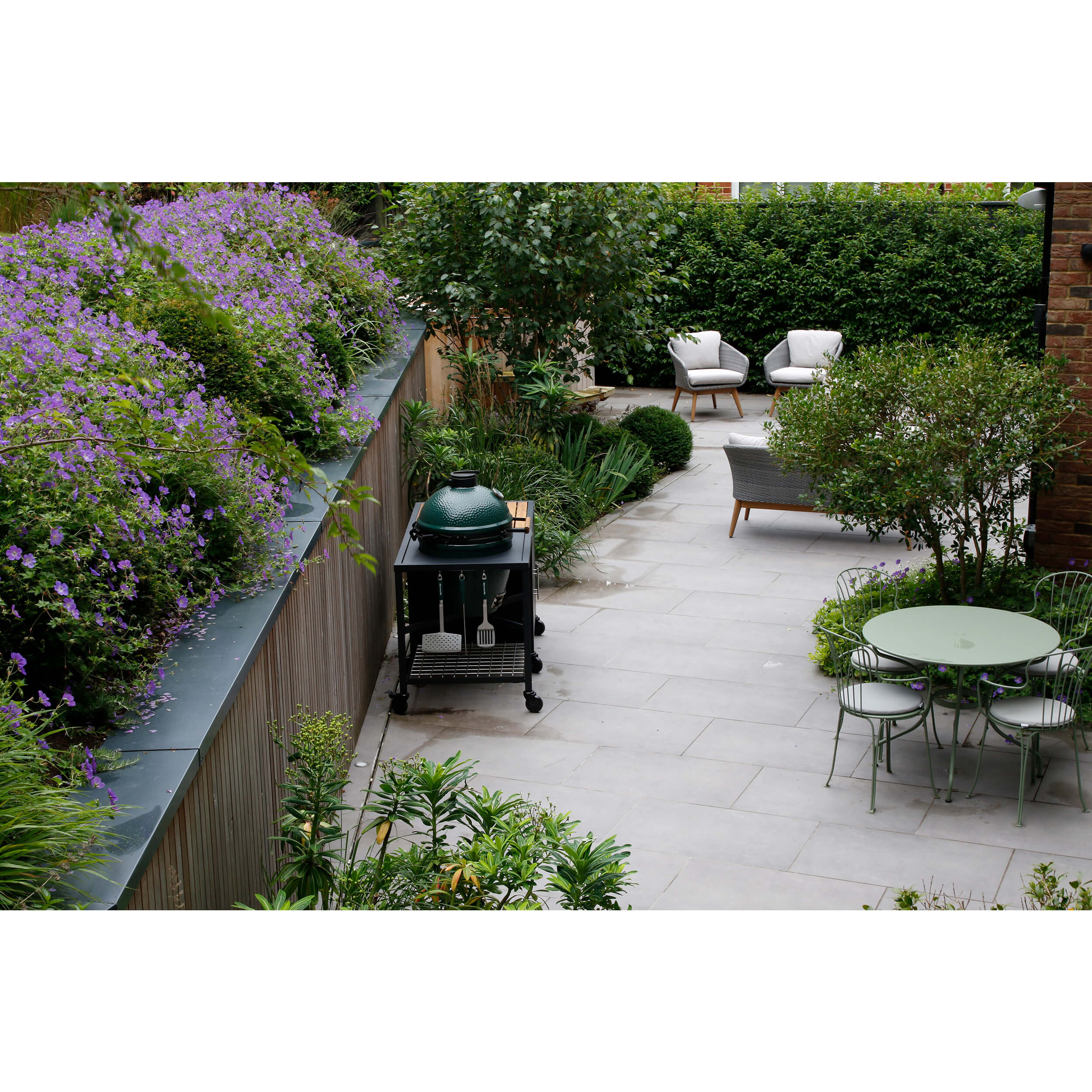 A garden cut into the hillside with a big green egg barbecue, green fermob table and chairs and comfy grey sofas