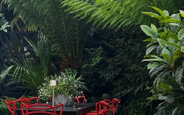 Outdoor dining table with bright red chairs among lush tropical plants and a 'TROPICAL VIBES' sign above.