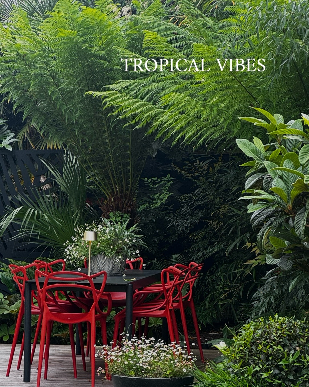 Outdoor dining table with bright red chairs among lush tropical plants and a 'TROPICAL VIBES' sign above.
