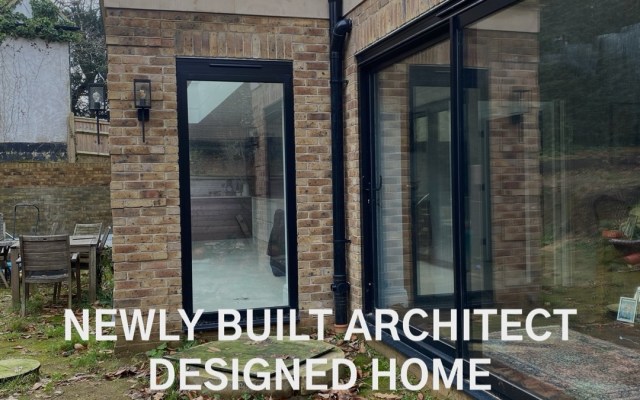Brick house exterior with large black-framed glass doors; leaves on the ground in a yard, overlay text reads 'New Project' and 'Newly Built Architect Designed Home'.